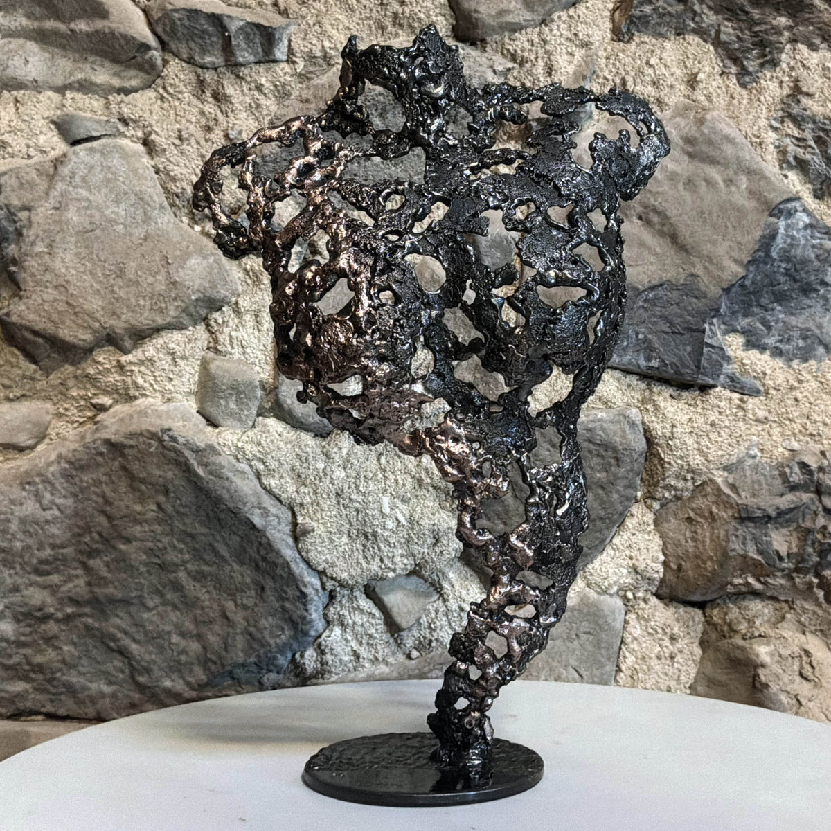 Les Traces du Temps, sculpture féminine en dentelle de métal présentée sur un meuble devant un mur en pierre dans un intérieur chaleureux.