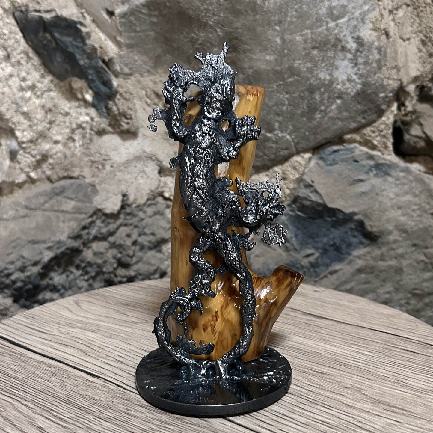 Sculpture contemporaine représentant une salamandre en dentelle de métal (acier), fixée sur un socle métallique circulaire et appuyée contre un morceau de bois naturel, œuvre de Philippe Buil, sur fond sombre.