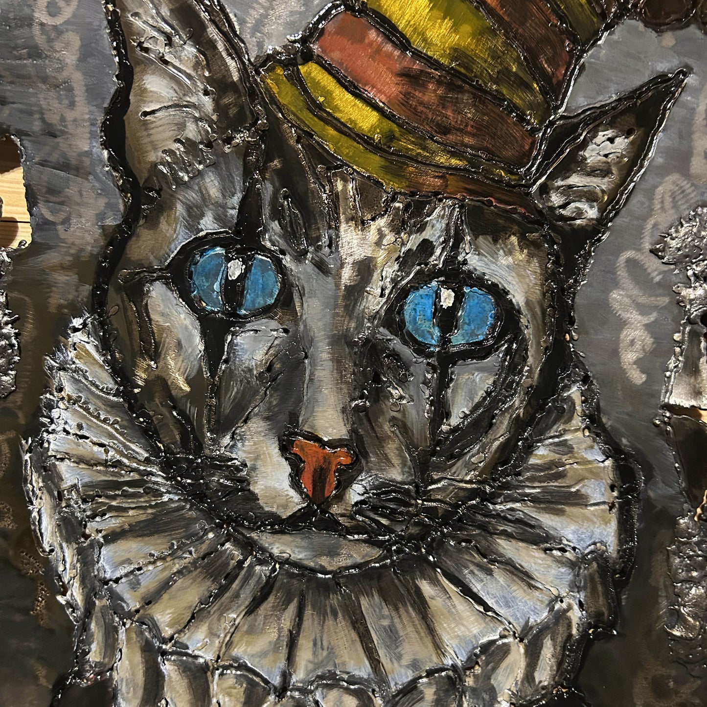 Sculpture murale de Philippe Buil sur tôle d’acier découpée, dentelle de métal et pigments : un chat arlequin expressif au regard bleu hypnotique.