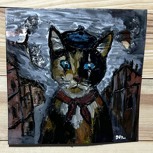 Tableau mural de 25×25 cm représentant un chat à béret dans un décor urbain gris et texturé. L’œuvre est réalisée en métal découpé avec des zones de dentelle de métal et colorée avec des pigments, créant un effet expressif et contrasté.
