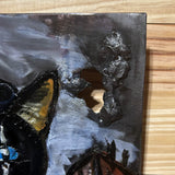 Tableau mural de 25×25 cm représentant un chat à béret dans un décor urbain gris et texturé. L’œuvre est réalisée en métal découpé avec des zones de dentelle de métal et colorée avec des pigments, créant un effet expressif et contrasté.