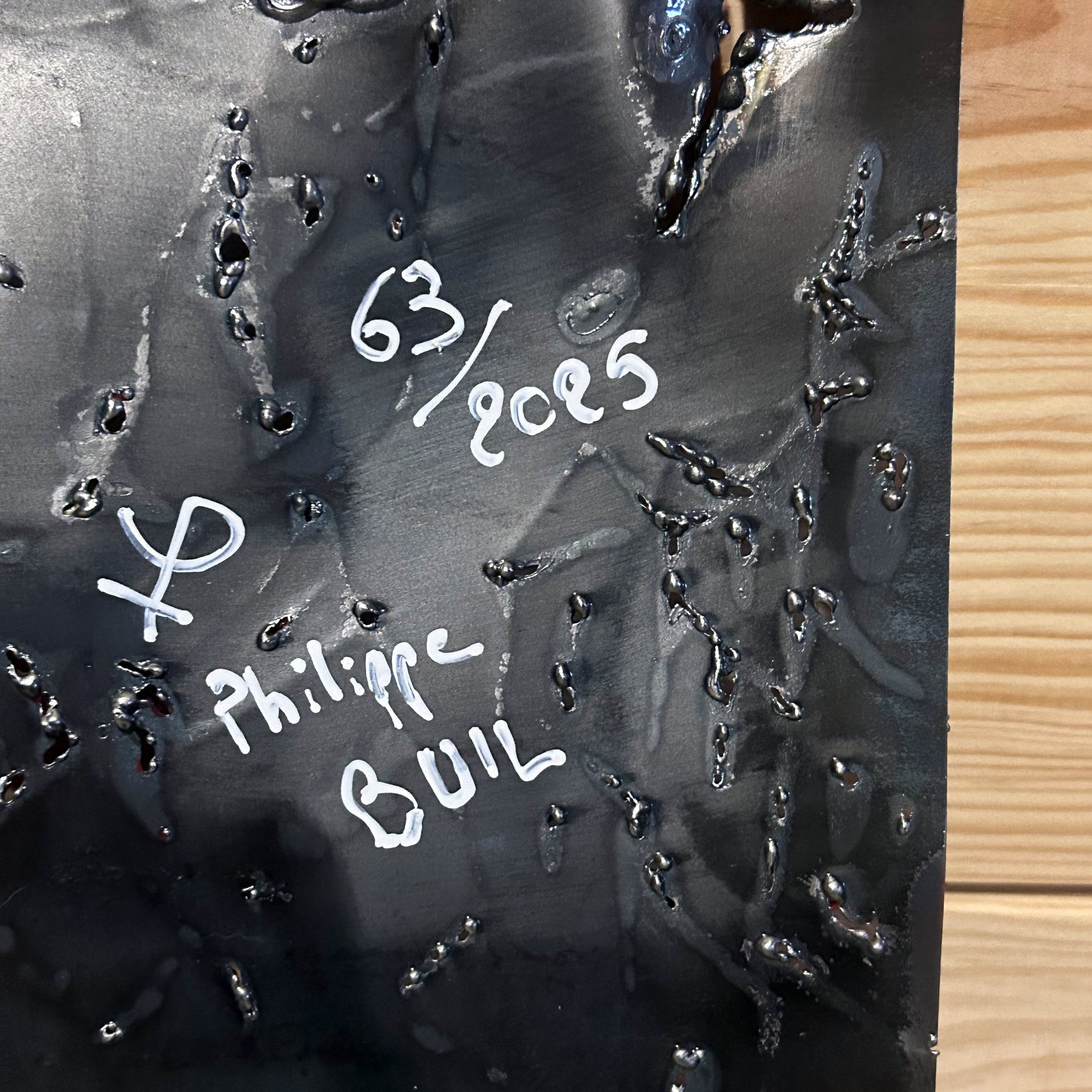 Tableau mural de 25×25 cm représentant un chat à béret dans un décor urbain gris et texturé. L’œuvre est réalisée en métal découpé avec des zones de dentelle de métal et colorée avec des pigments, créant un effet expressif et contrasté.