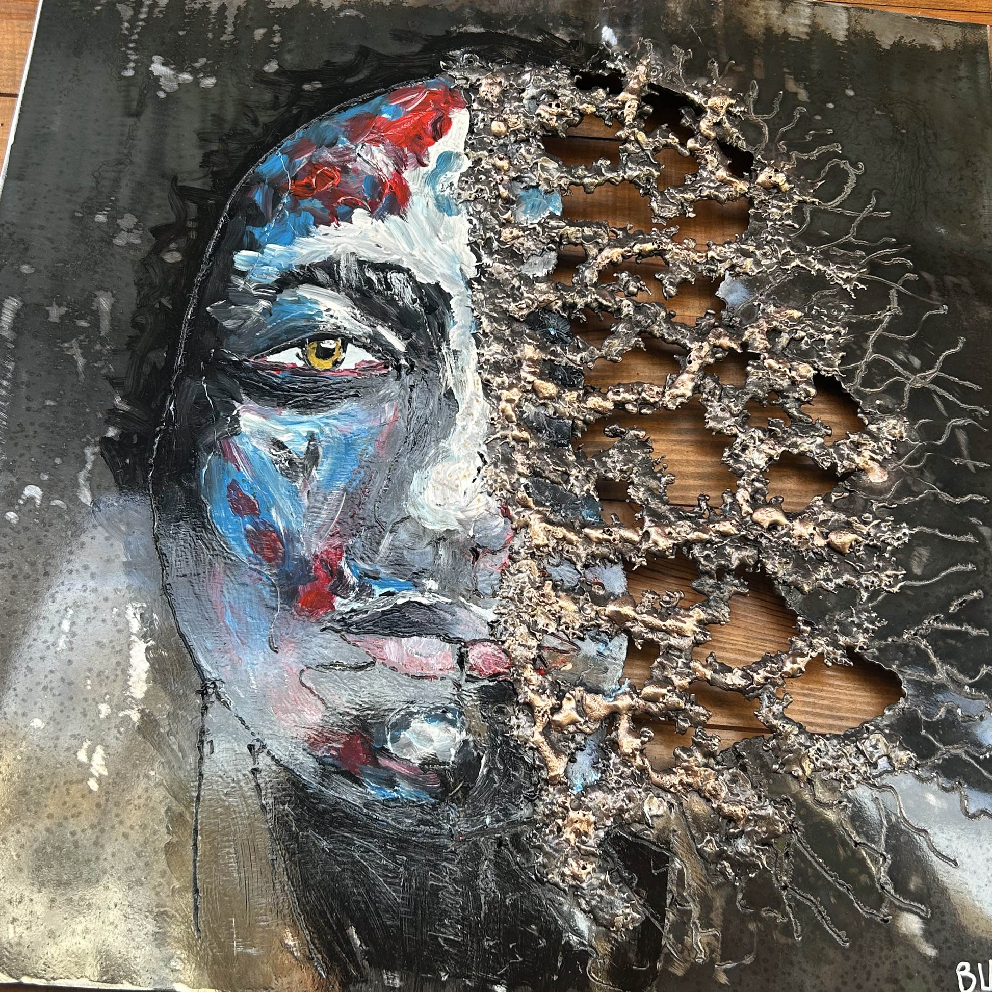 Tableau contemporain en métal de Philippe Buil représentant un visage en relief, moitié peint avec des pigments, moitié ajouré en dentelle d’acier et de bronze, créant un jeu d’ombres et de lumière.
