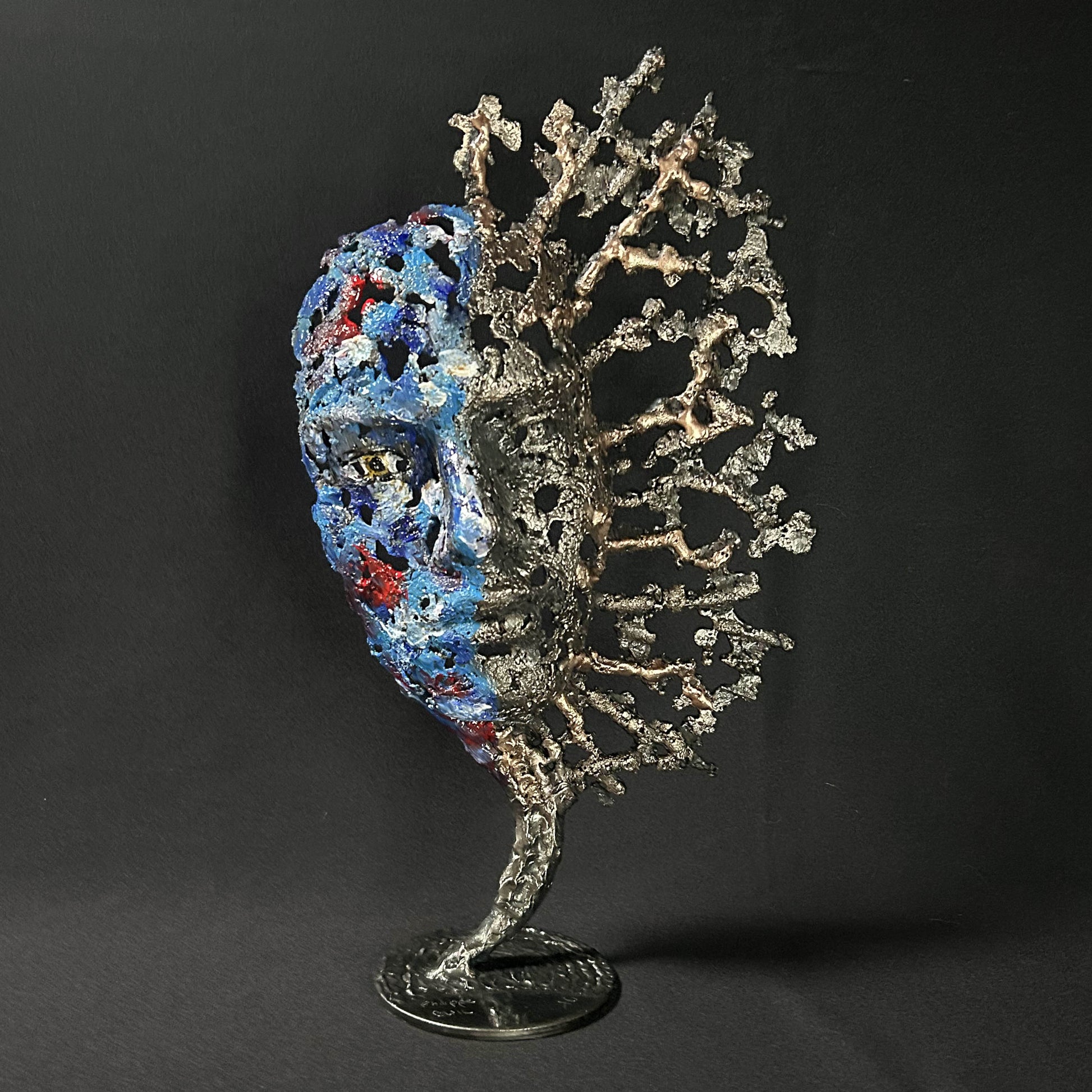 Sculpture de Philippe Buil représentant un visage humain en dentelle d'acier et bronze. Le côté gauche du visage est coloré avec des pigments bleus et rouges, tandis que le côté droit se transforme en une structure métallique ajourée et arborescente.