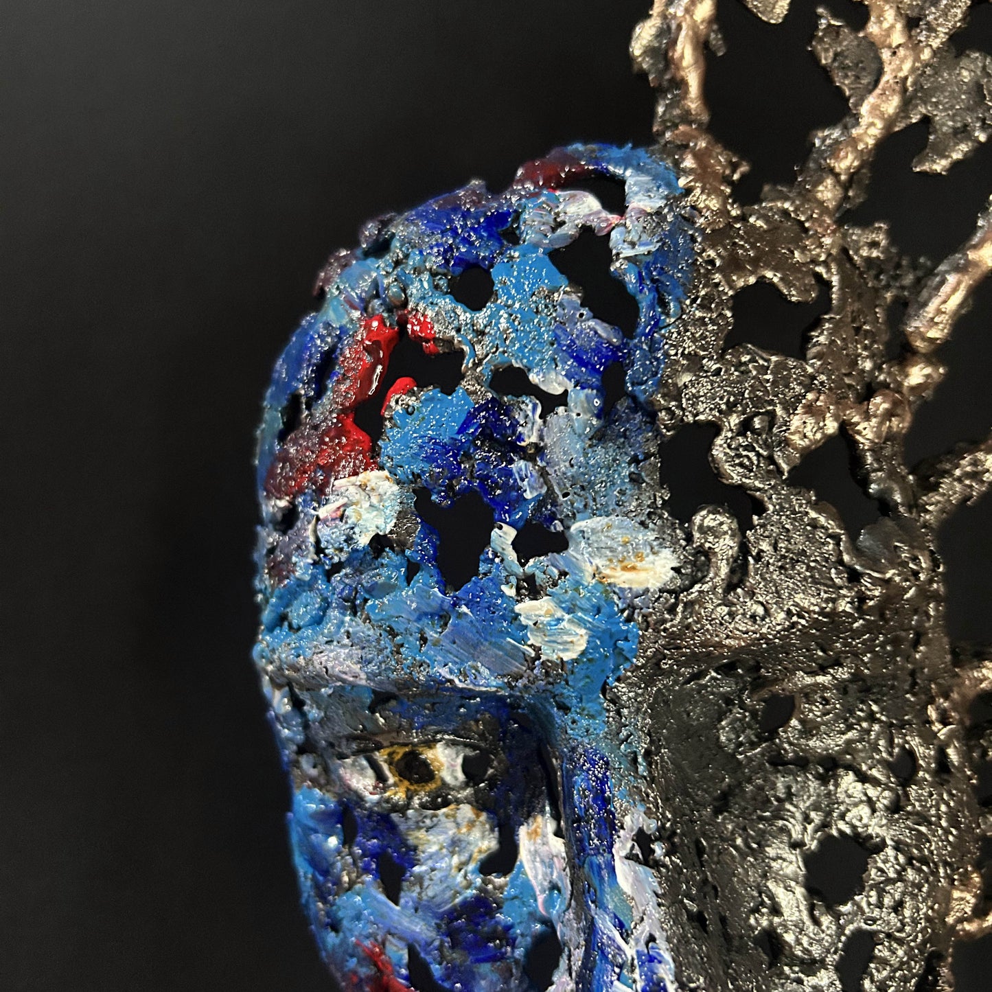 Sculpture de Philippe Buil représentant un visage humain en dentelle d'acier et bronze. Le côté gauche du visage est coloré avec des pigments bleus et rouges, tandis que le côté droit se transforme en une structure métallique ajourée et arborescente.