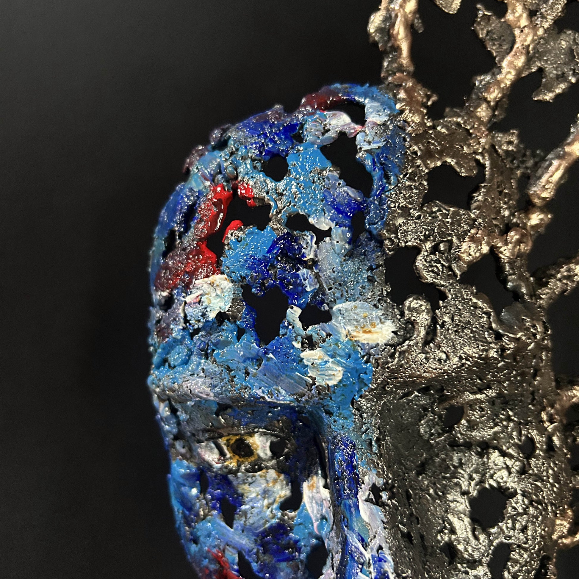 Sculpture de Philippe Buil représentant un visage humain en dentelle d'acier et bronze. Le côté gauche du visage est coloré avec des pigments bleus et rouges, tandis que le côté droit se transforme en une structure métallique ajourée et arborescente.