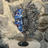 Sculpture de Philippe Buil représentant un visage humain en dentelle d'acier et bronze. Le côté gauche du visage est coloré avec des pigments bleus et rouges, tandis que le côté droit se transforme en une structure métallique ajourée et arborescente.