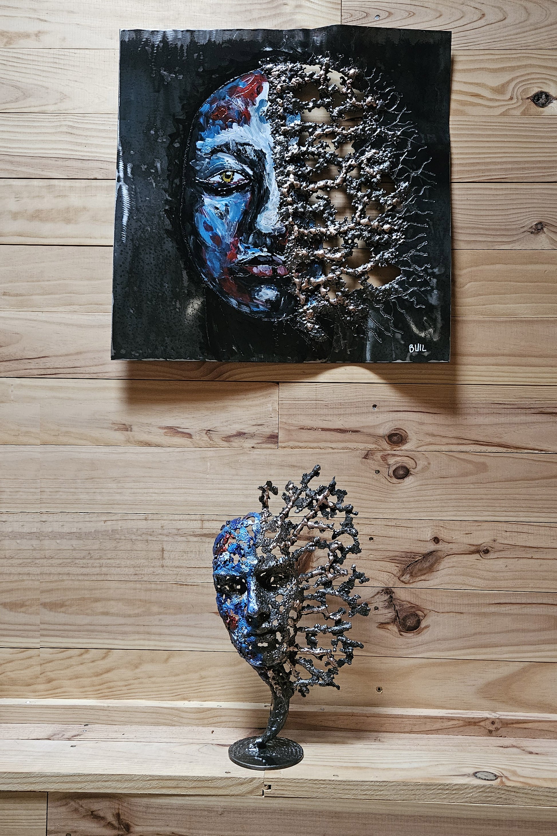 Sculpture de Philippe Buil représentant un visage humain en dentelle d'acier et bronze. Le côté gauche du visage est coloré avec des pigments bleus et rouges, tandis que le côté droit se transforme en une structure métallique ajourée et arborescente.