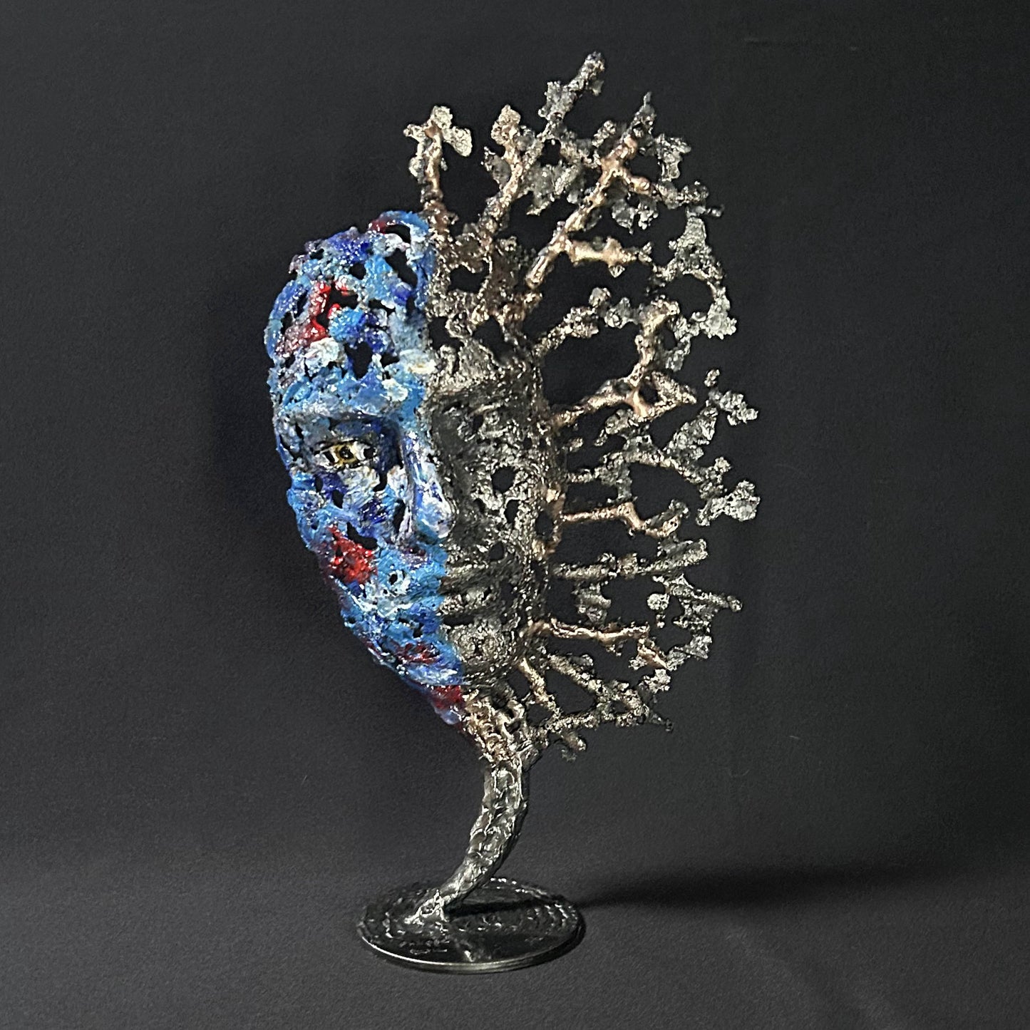 Sculpture de Philippe Buil représentant un visage humain en dentelle d'acier et bronze. Le côté gauche du visage est coloré avec des pigments bleus et rouges, tandis que le côté droit se transforme en une structure métallique ajourée et arborescente.