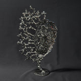 Sculpture de Philippe Buil représentant un visage humain en dentelle d'acier et bronze. Le côté gauche du visage est coloré avec des pigments bleus et rouges, tandis que le côté droit se transforme en une structure métallique ajourée et arborescente.