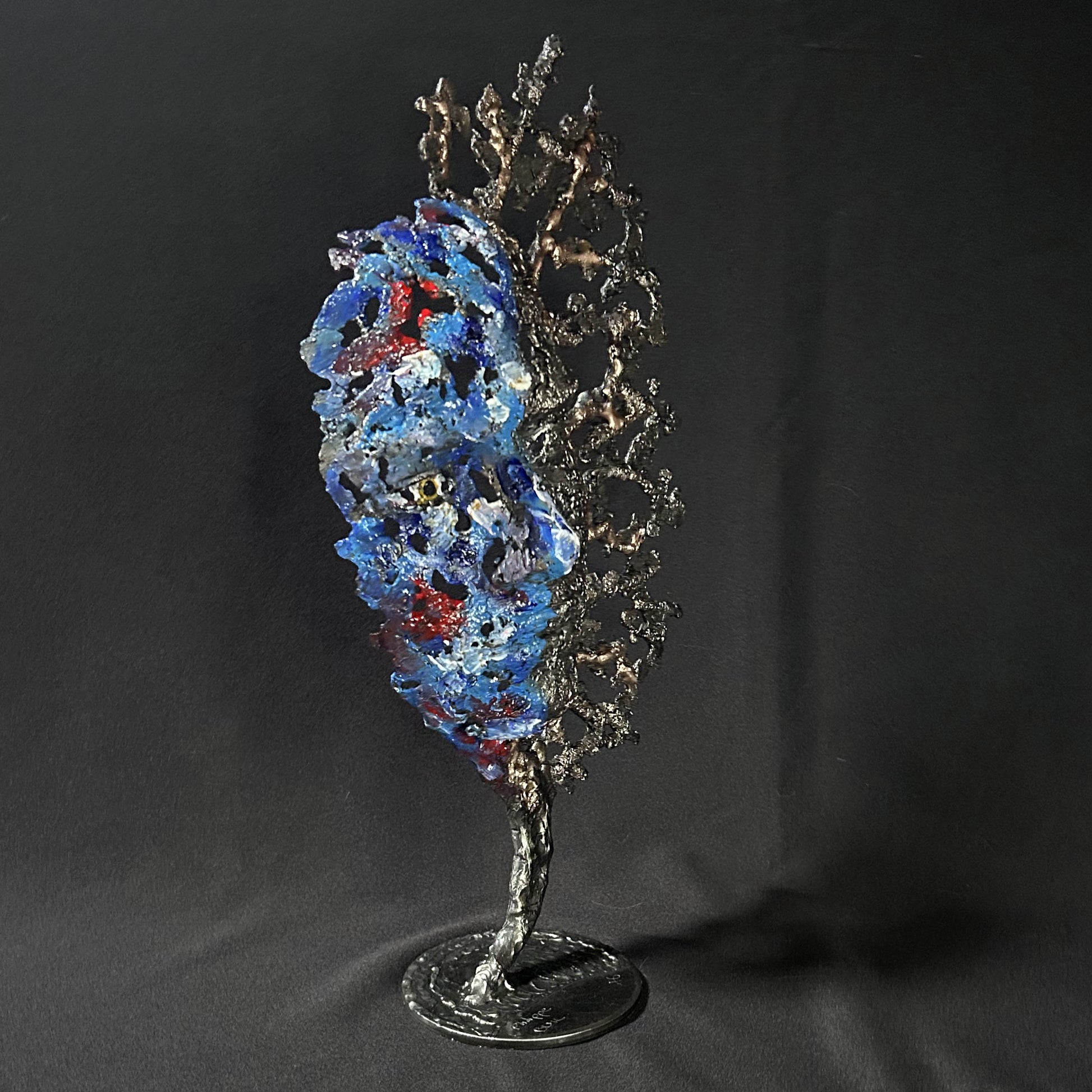 Sculpture de Philippe Buil représentant un visage humain en dentelle d'acier et bronze. Le côté gauche du visage est coloré avec des pigments bleus et rouges, tandis que le côté droit se transforme en une structure métallique ajourée et arborescente.
