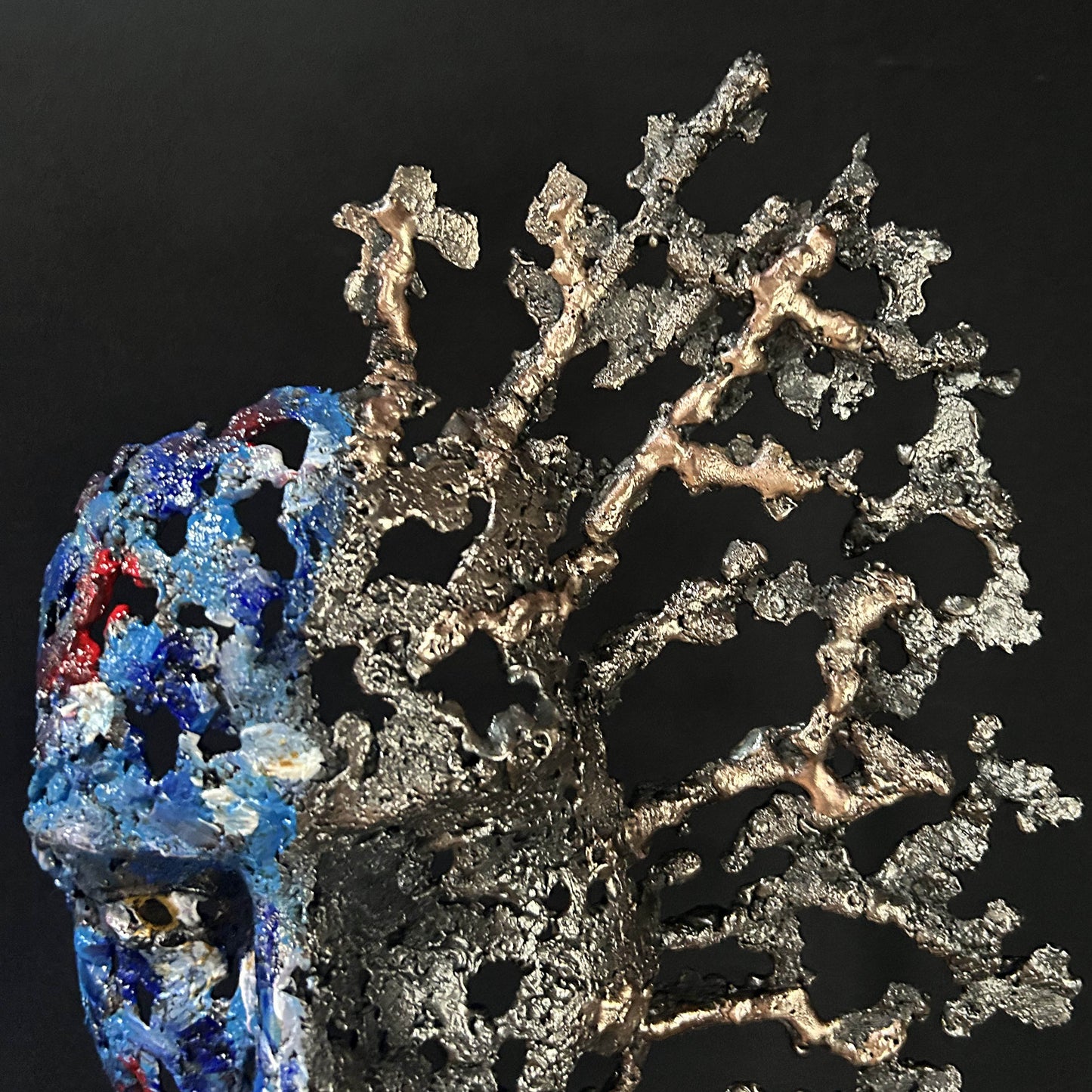 Sculpture de Philippe Buil représentant un visage humain en dentelle d'acier et bronze. Le côté gauche du visage est coloré avec des pigments bleus et rouges, tandis que le côté droit se transforme en une structure métallique ajourée et arborescente.