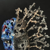 Sculpture de Philippe Buil représentant un visage humain en dentelle d'acier et bronze. Le côté gauche du visage est coloré avec des pigments bleus et rouges, tandis que le côté droit se transforme en une structure métallique ajourée et arborescente.