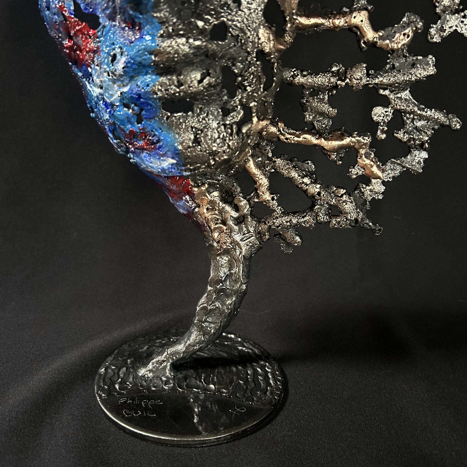 Sculpture de Philippe Buil représentant un visage humain en dentelle d'acier et bronze. Le côté gauche du visage est coloré avec des pigments bleus et rouges, tandis que le côté droit se transforme en une structure métallique ajourée et arborescente.