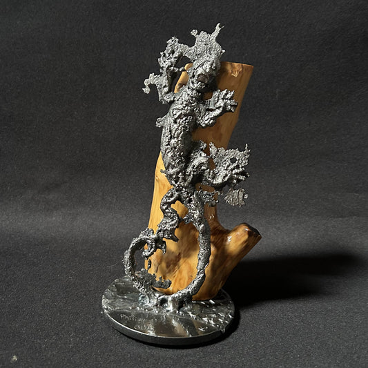 Sculpture contemporaine représentant une salamandre en dentelle de métal (acier), fixée sur un socle métallique circulaire et appuyée contre un morceau de bois naturel, œuvre de Philippe Buil, sur fond sombre.