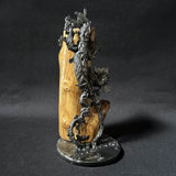 Sculpture contemporaine représentant une salamandre en dentelle de métal (acier), fixée sur un socle métallique circulaire et appuyée contre un morceau de bois naturel, œuvre de Philippe Buil, sur fond sombre.